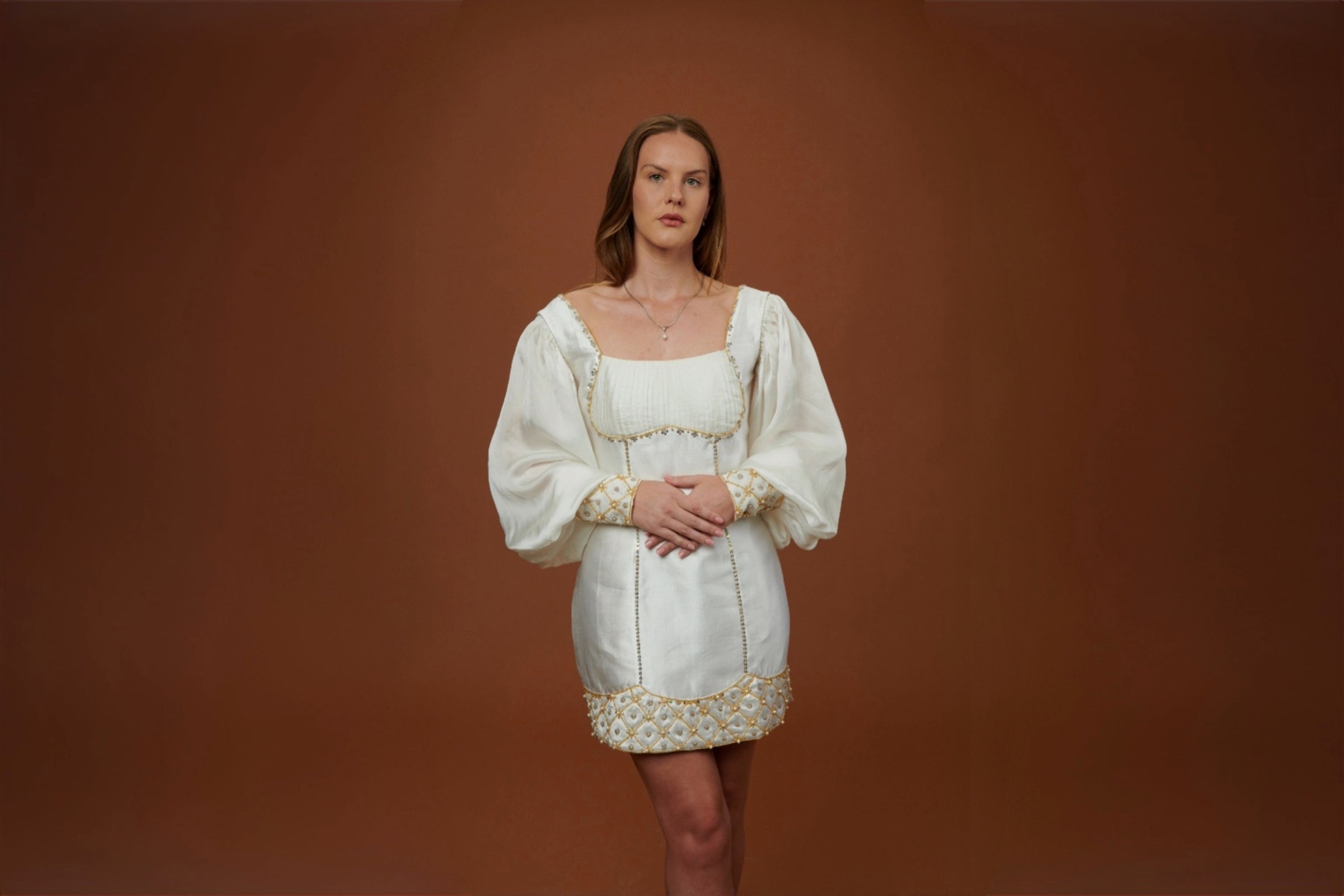 An ivory beaded dress in a composed standing pose, against a wide clay backdrop.